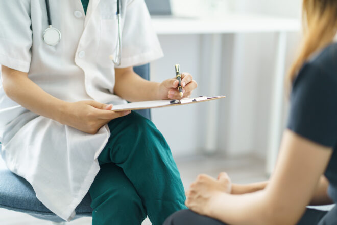 Zdjęcie lekarki, która trzyma w dłoni długopis i papiery. Naprzeciwko siedzi pacjentka.