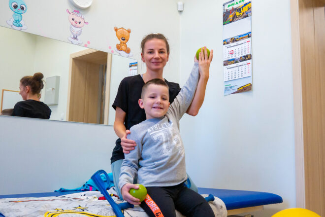 Zdjęcie rehabilitantki, która trenuje z chłopcem. Chłopiec trzyma rękę w górze, obok leży żółta piłka i kula.