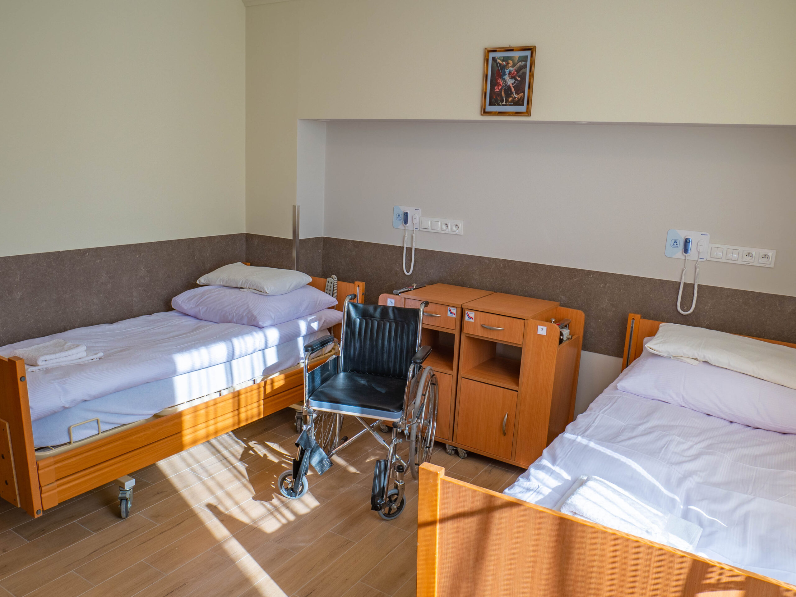 Zdjęcie pokoju w Warszawskim domu seniora. Na zdjęciu widać dwa łóżka, dwie szafki, wózek inwalidzki i telefony do wzywania obsługi.
