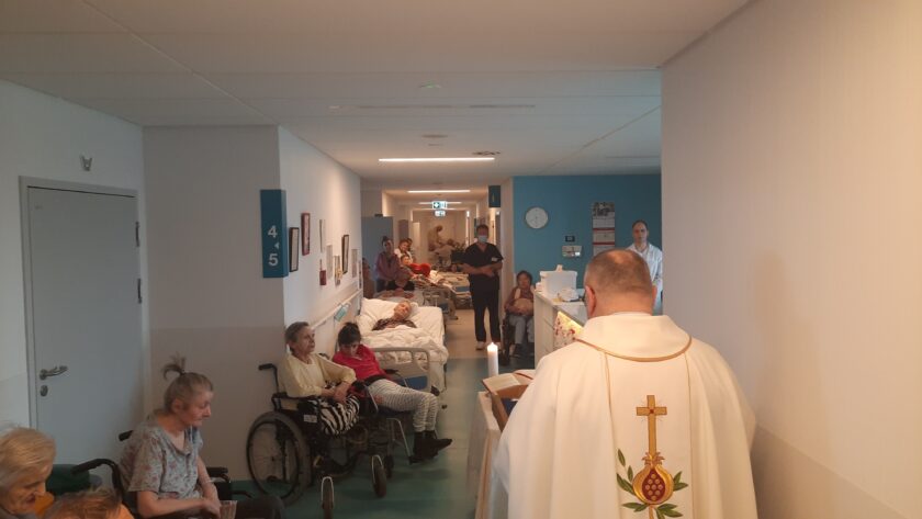 Zdjęcie księdza na korytarzu w szpitalu. Na zdjęciu znajdują się również pacjenci na wózkach inwalidzkich i łóżkach.
