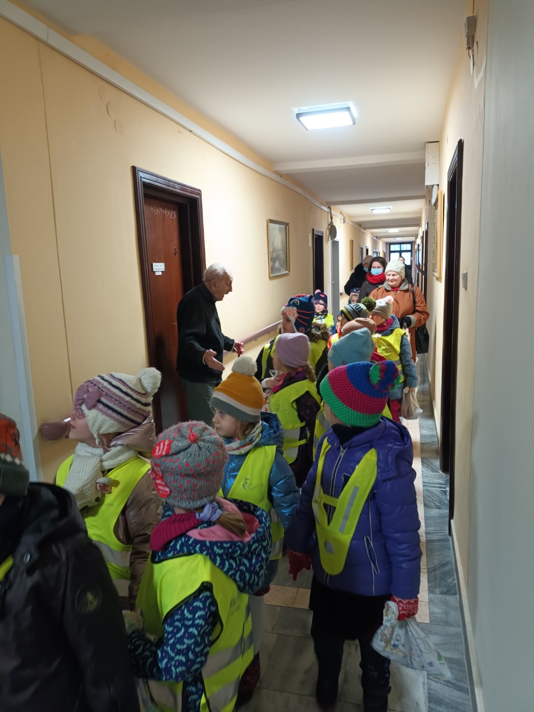 Zdjęcie przedszkolaków ubranych w odblaskowe kamizelki, które przyszły do domu seniora. Na zdjęciu widać też seniora, który cieszy się na widok dzieci.