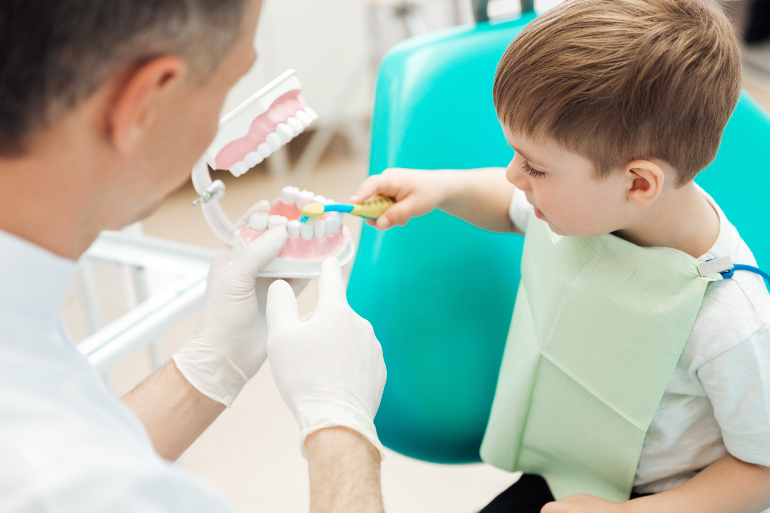 Zdjęcie dentysty, który trzyma w dłoni sztuczną szczękę. Naprzeciwko niego stoi chłopiec, który szczoteczką do zębów uczy się myć zęby.