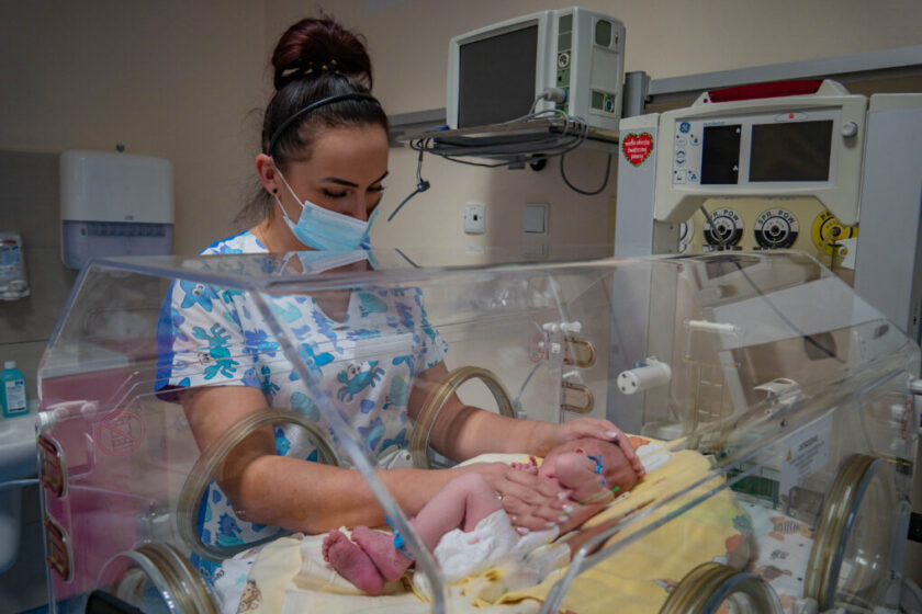 Zdjęcie położnej, która stoi obok inkubatora, w którym jest noworodek.