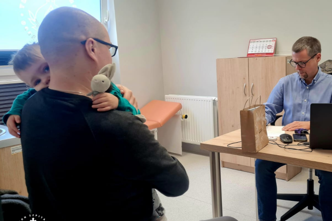 Fotografia przedstawia dwóch mężczyzn (jednego z dzieckiem na ramieniu) siedzących w gabinecie lekarskim