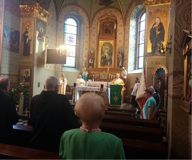 Fotografia przedstawia uroczystość mszy świętej w kościele, w której uczestniczy grupa osób. Na ołtarzu widać księdza przemawiającego przez mikrofon oraz stojących obok, dwóch innych księży modlących się. Przed ołtarzem widać inne osoby uczestniczące we mszy świętej.