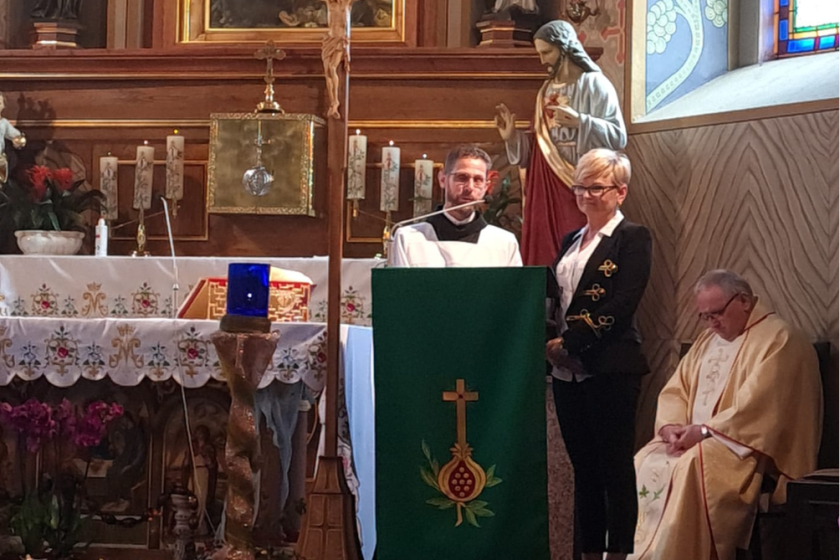 Fotografia przedstawia zakonnika przemawiającego przez mikrofon na ołtarzu w kościele. Obok zakonnika stoi elegancko ubrana kobieta, obok na krześle siedzi ksiądz.