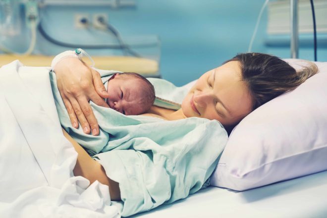 Zdjęcie przedstawia młodą kobietę leżącą na szpitalnym łóżku, trzymającą na piersi noworodka