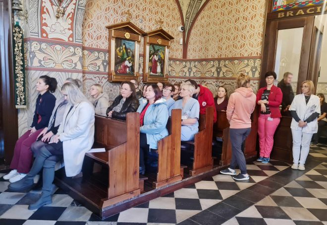 Fotografia przedstawia grupę osób siedzących w kościelnych ławkach podczas mszy świętej. W tle widać osoby stojące.