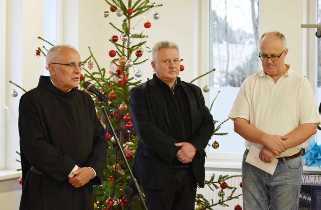 Fotografia przedstawia trzech mężczyzn stojących obok mikrofonu, jeden z nich - brat Bonifrater przemawia przez mikrofon. W tle stoi świąteczna choinka.