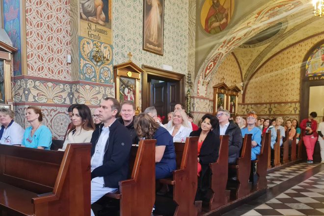 Fotografia przedstawia grupę osób siedzących w kościelnych ławkach podczas mszy świętej. W tle widać osoby stojące.