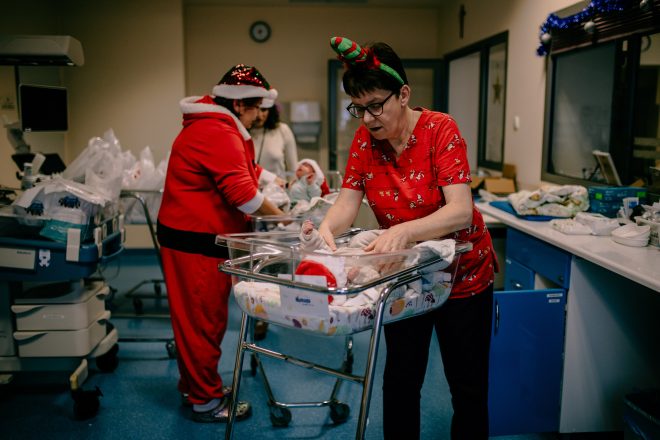 Fotografia przedstawia kobiety ubrane w czerwone, świąteczne stroje, które zajmują się noworodkami w sali szpitalnej