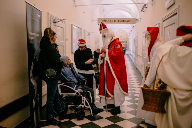 Zdjęcie przedstawia grupę osób stojących na korytarzu szpitalnym. Wśród nich są trzy osoby w strojach nawiązujących do Świąt Bożego Narodzenia oraz mężczyzna w stroju Świętego Mikołaja. Obok i w tle widać pacjentów.