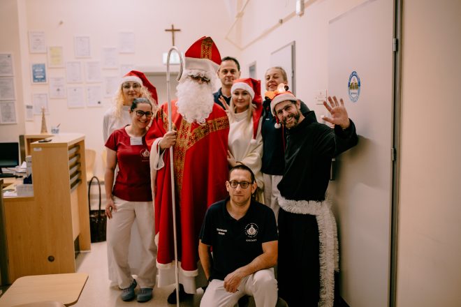 Zdjęcie przedstawia grupę uśmiechających się osób pozujących do zdjęcia z mężczyzną przebranym za świętego Mikołaja. Dwie kobiety posiadają świąteczne nakrycia głowy - czapki Mikołaja. Zdjęcie zostało wykonane w holu budynku szpitala