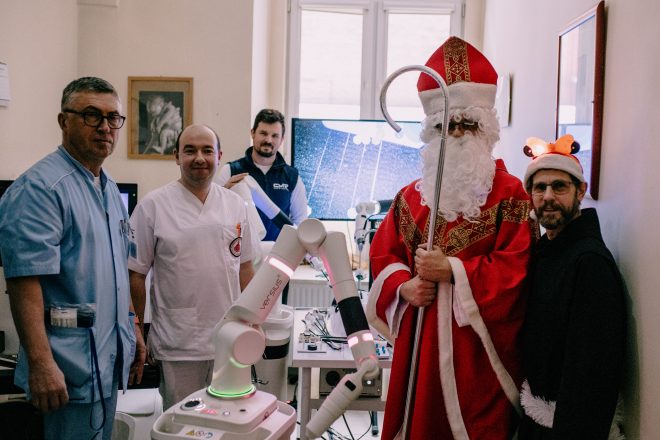 Zdjęcie przedstawia grupę uśmiechających się mężczyzn w strojach lekarskich pozujących do zdjęcia z mężczyzną w stroju Świętego Mikołaja. Wśród osób jest również mężczyzna w stroju zakonnika. Zdjęcie został zrobione w gabinecie lekarskim.