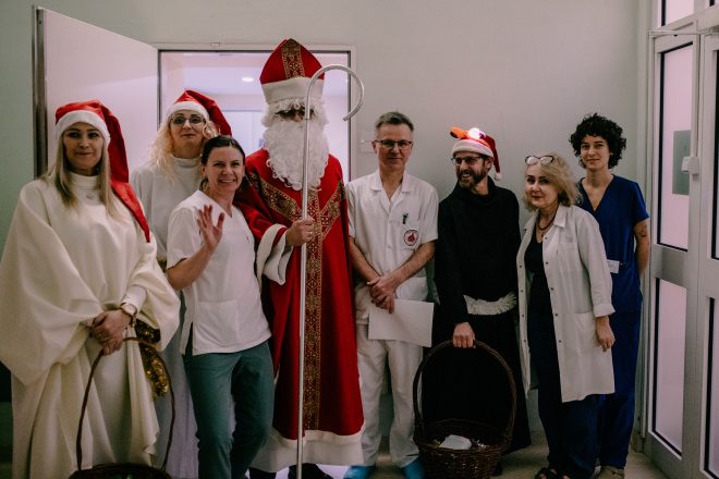 Fotografia przedstawia grupę osób, pięć kobiet, dwóch mężczyzn i jednego mężczyznę przebranego w strój Świętego Mikołaja. Wszystkie osoby uśmiechają się, dwie kobiety i jeden mężczyzna - w czarnym habicie, mają na głowach czapki Mikołaja. Wszystkie osoby pozują do zdjęcia w pomieszczeniu szpitalnym