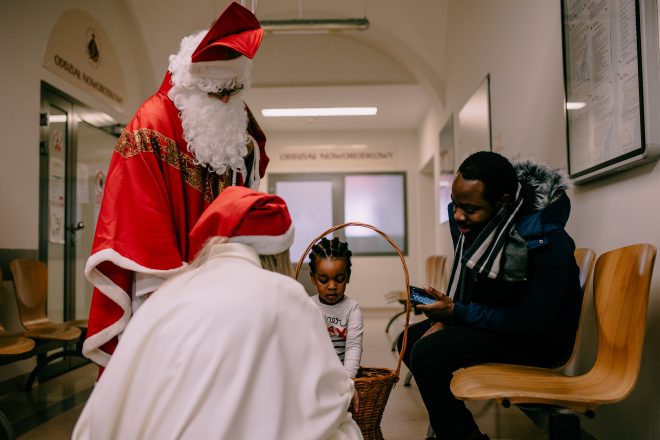 Fotografia przedstawia mężczyznę w stroju Świętego Mikołaja, który razem z kobietą w czerwonej czapce Mikołaja, rozmawia z małą dziewczynką i mężczyzną w korytarzu szpitalnym