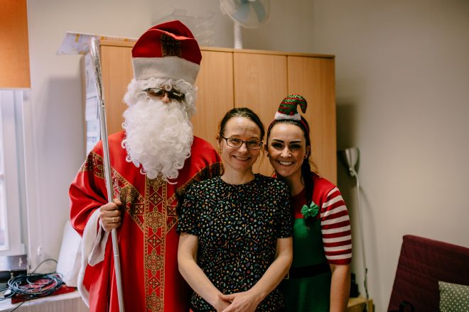 Zdjęcie przedstawia dwie uśmiechające się kobiety, z których jedna ma strój nawiązujący do Świąt Bożego Narodzenia. Obok kobiet stoi mężczyzna w stroju Świętego Mikołaja. Zdjęcie zostało wykonane w pomieszczeniu zamkniętym.
