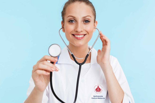 Zdjęcie przedstawia uśmiechniętą, młoda kobietę lekarza, stojącą na niebieskim tle, trzymającą w rękach stetoskop