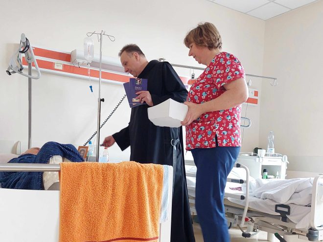 Zdjęcie obrazuje brata bonifratra, który stoi wraz z pielęgniarką przy szpitalnym łóżku z pacjentem