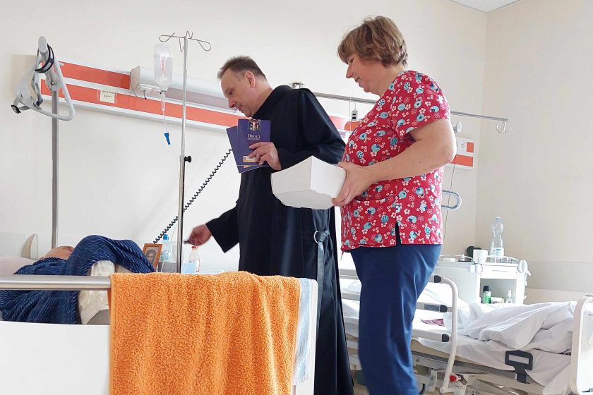 Zdjęcie obrazuje brata bonifratra, który stoi wraz z pielęgniarką przy szpitalnym łóżku z pacjentem
