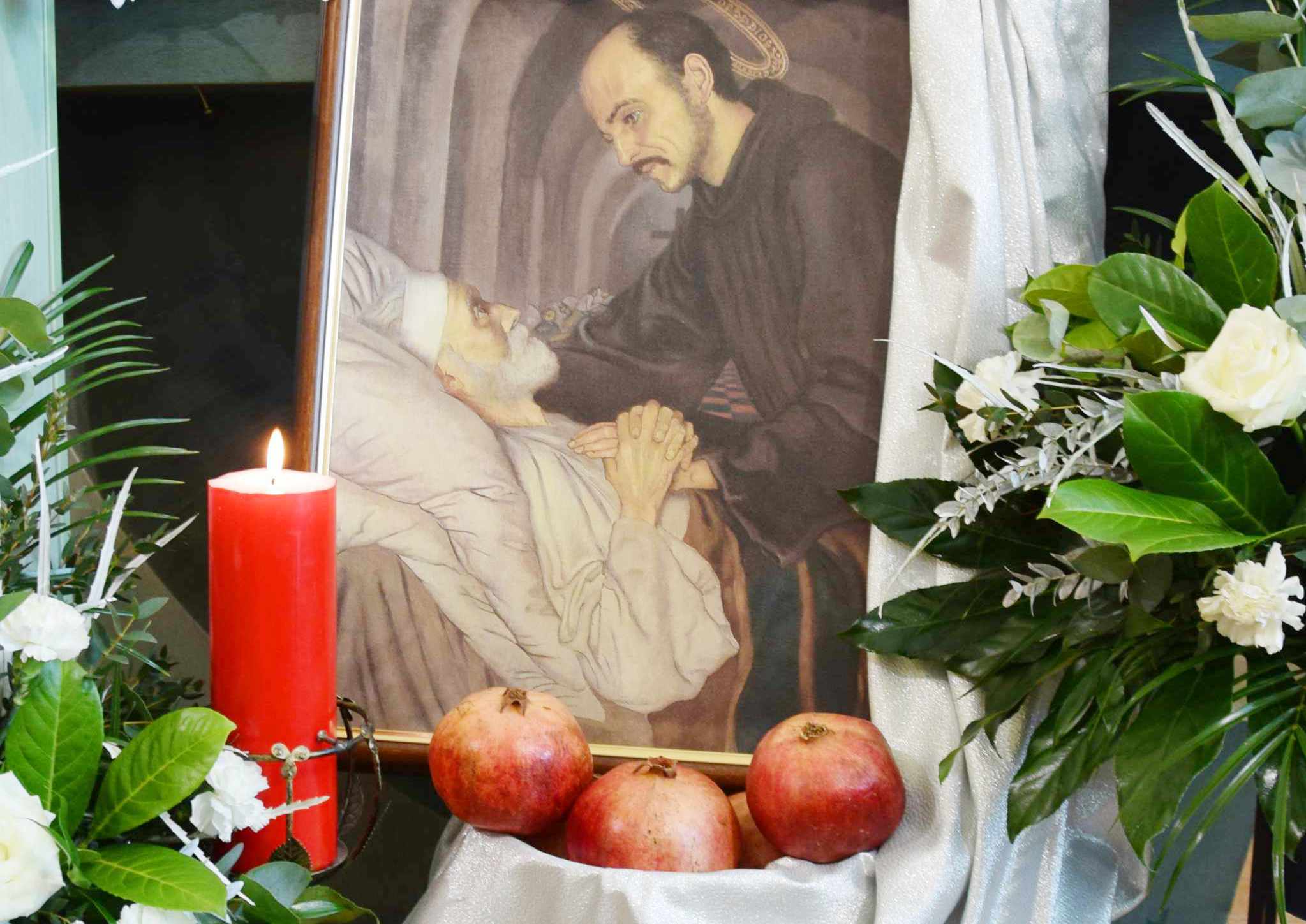 Zdjęcie przedstawia obraz z postacią świętego Jana Bożego pochylającego się nad chorym, leżącym człowiekiem. Obok obrazu widać kwiaty, świecę oraz owoce granatu