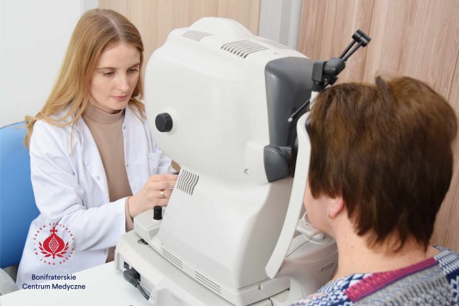 Zdjęcie przedstawia lekarza okulistę siedzącego przed dużym urządzeniem diagnostycznym. Z drugiej strony urządzenia siedzi pacjentka, która spogląda w specjalny okular. Fotografia została wykonana w gabinecie lekarskim.