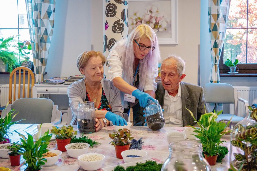 Zdjęcie przedstawia dwójkę seniorów - kobietę i mężczyznę uczestniczących sadzących kwiatki wspólnie z młodą kobietą - opiekunką. Osoby te znajdują się w pomieszczeniu zamkniętym i siedzą przy dużym stole, na którym widać rośliny oraz dekoracje
