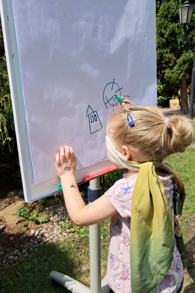 Zdjęcie przedstawia młodą dziewczynkę malującą mazakiem na dużej, pionowej kartce umieszczonej na sztaludze stojącej w ogrodzie