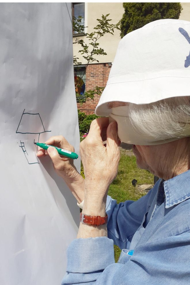 Zdjęcie przedstawia starszą kobietę rysującą na dużej, pionowej kartce