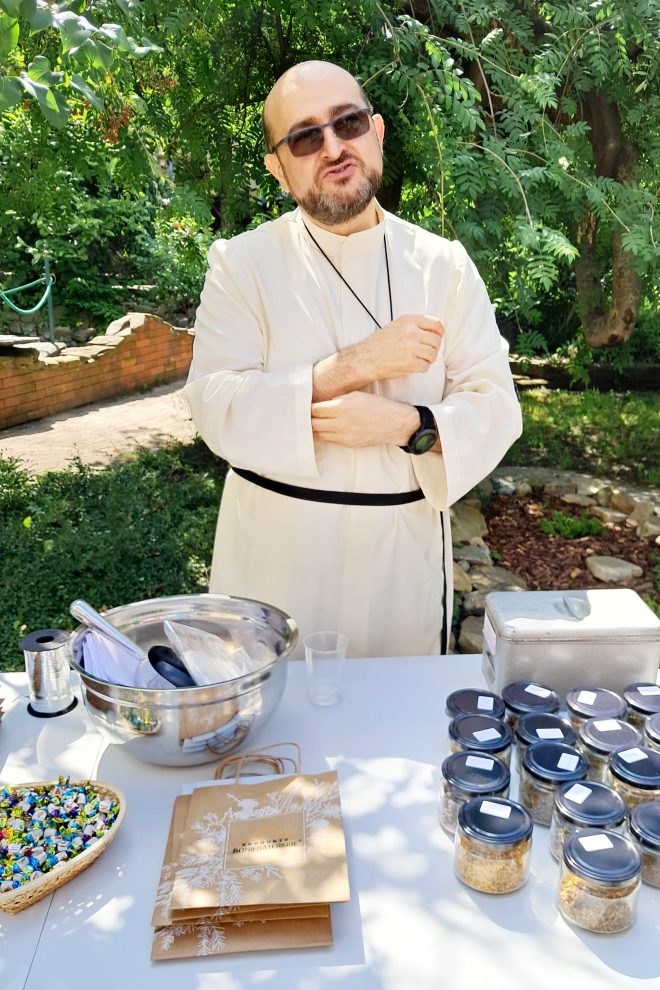 Zdjęcie przedstawia zakonnika stojącego obok stołu na którym umieszczone są zioła w słoikach, cukierki oraz torebki papierowe