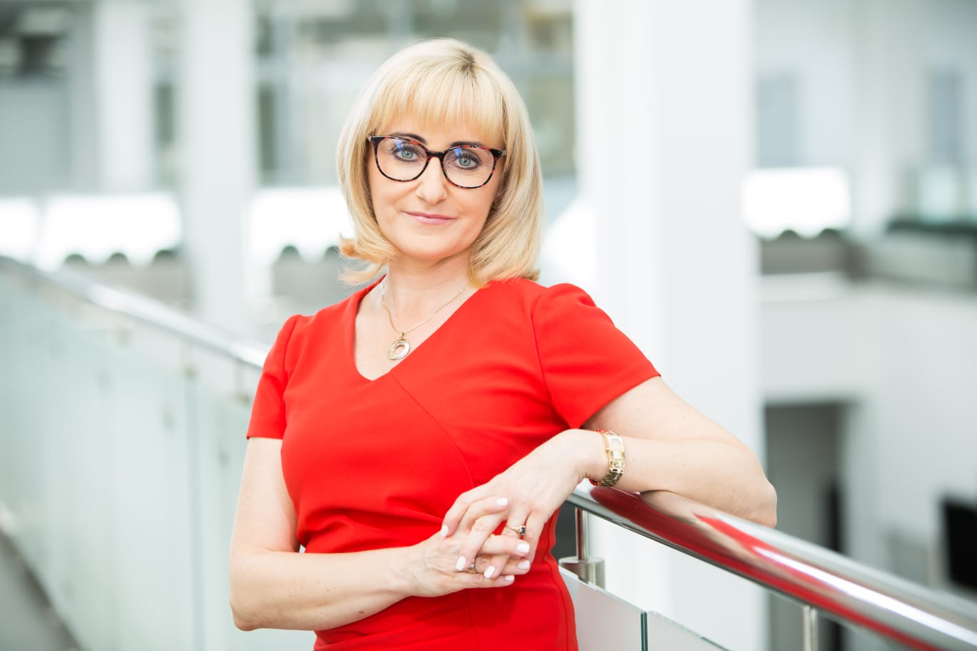 Zdjęcie przedstawia elegancko ubraną kobietę, uśmiechającą się do obiektywu aparatu, opierającą się o balustradę