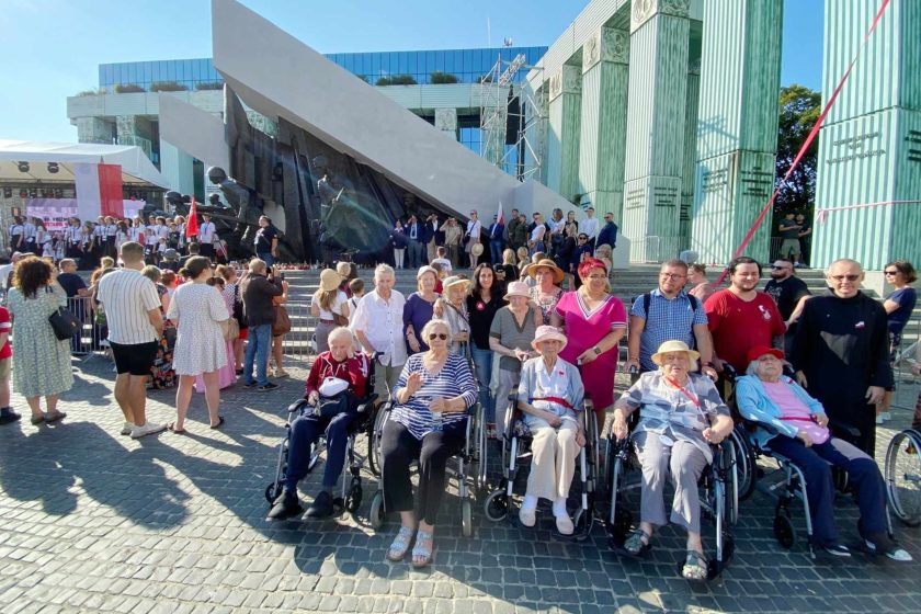 Fotografia przedstawia grupę osób na wózkach inwalidzkich obok których stoją osoby pełnosprawne, przed pomnikiem Powstańców Warszawskich