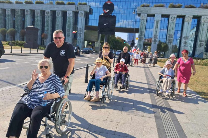 Fotografia przedstawia grupę osób poruszających się na wózkach inwalidzkich, którym pomagają opiekunowie, przemieszczającą się chodnikiem w mieście