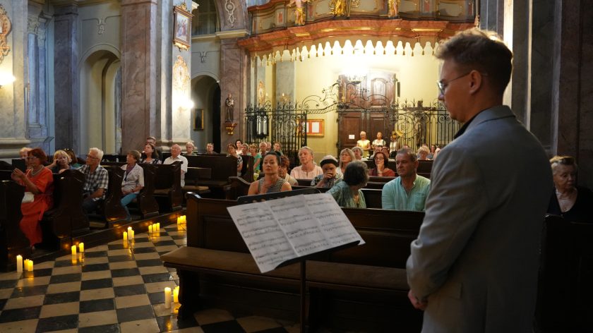 Zdjęcie przedstawia scenę wewnątrz kościoła, w której grupa ludzi siedzi w ławkach, zwrócona w stronę mężczyzny stojącego tyłem, ubranym w elegancki garnitur. Przed nim stoi pulpit z nutami. W tle widać ozdobny ołtarz, marmurowe filary oraz dekoracyjne elementy architektoniczne. Podłoga jest wyłożona czarno-białymi płytkami, a na ziemi wokół ławek rozstawione są zapalone świece, co tworzy nastrojową atmosferę.