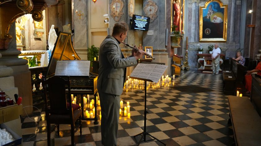 Mężczyzna w szarym garniturze gra na flecie poprzecznym wewnątrz kościoła. Przed nim stoi pulpit z nutami, a w tle widoczny jest zabytkowy instrument klawiszowy, otoczony licznymi zapalonymi świecami. Wnętrze kościoła jest bogato zdobione, z widocznymi rzeźbami, obrazami i marmurowymi elementami architektonicznymi. Na ścianie po prawej stronie wisi obraz przedstawiający papieża, a na podłodze ułożone są czarno-białe płytki. Atmosfera jest podniosła i skupiona na występie muzycznym.