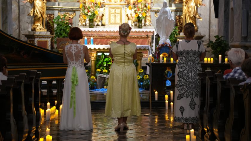 Na zdjęciu widzimy trzy kobiety stojące tyłem do obiektywu w kościele, zwrócone w stronę ołtarza. Kobiety mają na sobie eleganckie, długie sukienki: pierwsza z lewej w białej sukni z koronkowym tyłem, środkowa w żółtej sukience z kwiatami we włosach, a trzecia w sukience z czarno-białym, ozdobnym wzorem. Przed nimi znajduje się ołtarz udekorowany kwiatami, świecami i złotymi elementami. Podłoga pokryta jest płytkami, a po bokach widać zapalone świece oraz siedzących wiernych.