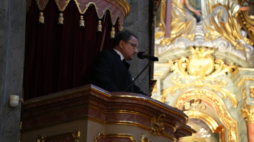 Mężczyzna w ciemnym garniturze stoi przy mównicy w kościele, przemawiając do mikrofonu. Mównica jest ozdobiona złotymi elementami i aksamitną zasłoną w kolorze bordowym. W tle widać fragment bogato zdobionego ołtarza z elementami złota i rzeźbami. Mężczyzna jest zwrócony w profil, a jego twarz wyraża skupienie.