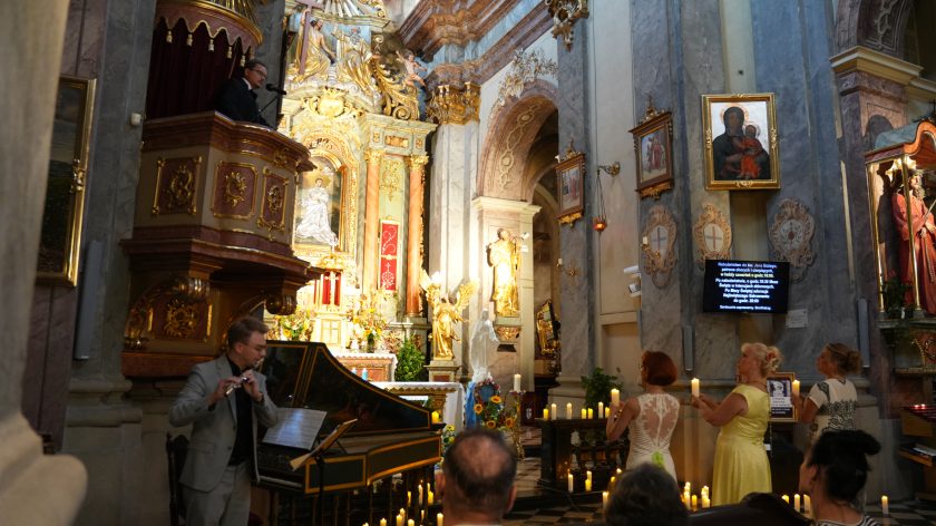 Na zdjęciu widzimy wnętrze bogato zdobionego kościoła podczas uroczystości. W lewej części kadru, na ambonie ozdobionej złotymi elementami, stoi mężczyzna w ciemnym garniturze, przemawiający do mikrofonu. Poniżej, po lewej stronie, mężczyzna w jasnym garniturze gra na flecie. Na środku kadru znajdują się trzy kobiety stojące tyłem do obiektywu, każda z nich trzyma zapaloną świecę. W tle widać złoty ołtarz, obrazy religijne oraz rzeźby. Na prawo od kobiet znajduje się ekran, na którym wyświetlone są informacje dotyczące nabożeństwa.