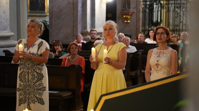 Na zdjęciu widzimy trzy kobiety stojące obok siebie w kościele, każda trzyma zapaloną świecę w dłoniach. Kobieta po lewej stronie ubrana jest w białą suknię z czarno-białym wzorem, środkowa w żółtą sukienkę i naszyjnik z koralików, a kobieta po prawej w białą suknię z koronkowymi detalami. Wszystkie są skupione na ceremonii. W tle widać innych uczestników siedzących w ławkach, obserwujących wydarzenia z uwagą.