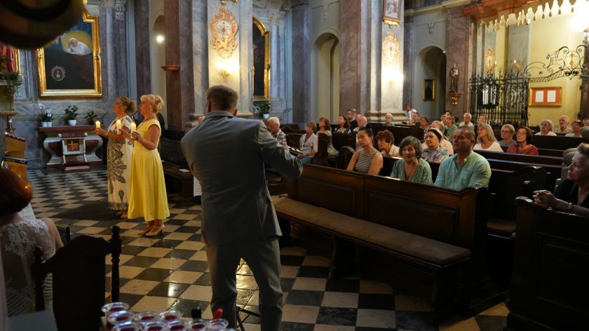 Na zdjęciu widzimy wnętrze kościoła podczas uroczystości. Na pierwszym planie po lewej stronie znajdują się dwie kobiety trzymające zapalone świece, ubrane w eleganckie suknie – jedna w białą suknię z czarnym wzorem, druga w żółtą sukienkę. Przed nimi stoi mężczyzna w szarym garniturze, który prawdopodobnie przewodniczy ceremonii. Po prawej stronie zdjęcia, w ławkach, siedzi grupa wiernych, którzy z uwagą obserwują wydarzenia. Na ścianach kościoła widoczne są obrazy i ozdobne detale architektoniczne.