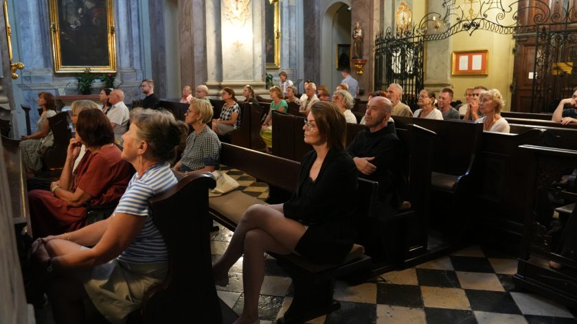 Na zdjęciu widzimy grupę ludzi siedzących w ławkach kościelnych, uważnie uczestniczących w uroczystości. W pierwszym planie, po prawej stronie, znajduje się kobieta w ciemnym ubraniu oraz mężczyzna ubrany w ciemną bluzę z kapturem, oboje siedzący w skupieniu. Reszta wiernych również siedzi w ławkach, patrząc w stronę ołtarza lub osoby prowadzącej ceremonie. W tle widoczne są obrazy na ścianach kościoła oraz ozdobne elementy architektoniczne.