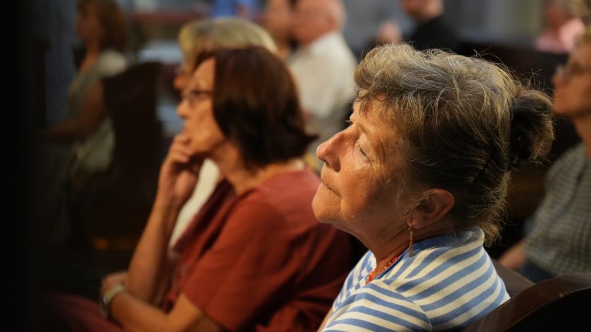 Na zdjęciu widzimy zbliżenie na twarz starszej kobiety siedzącej w ławce kościelnej. Kobieta ma siwe włosy upięte w kok i nosi koszulkę w biało-niebieskie paski. Jej wyraz twarzy sugeruje skupienie i zadumę podczas uczestnictwa w ceremonii. W tle, w nieostrości, widoczne są inne osoby, także siedzące w ławkach, pogrążone w modlitwie lub refleksji.