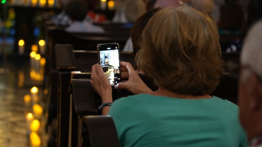 Na zdjęciu widzimy kobietę siedzącą w ławce kościelnej, która trzyma w rękach telefon komórkowy i robi zdjęcie wnętrza kościoła. Widok na ekranie telefonu pokazuje ołtarz i ławki w kościele. Kobieta ubrana jest w zieloną bluzkę, a jej krótkie, jasne włosy są widoczne od tyłu. W tle widać innych wiernych, którzy również siedzą w ławkach.