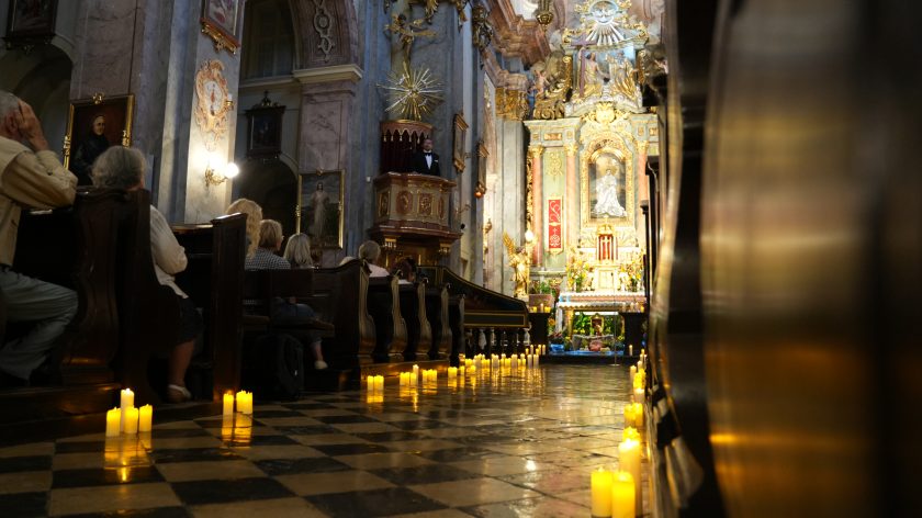 Zdjęcie przedstawia wnętrze bogato zdobionego kościoła podczas uroczystości. Widok skierowany jest w stronę ołtarza, który jest ozdobiony złotymi elementami, kwiatami i obrazami religijnymi. Po lewej stronie, na ambonie, przemawia mężczyzna w czarnym garniturze z muszką. Posadzka kościoła jest wyłożona szachownicą z ciemnych i jasnych płytek, na której ustawione są zapalone świece, dodające nastrojowego światła. W ławkach siedzą wierni, uważnie obserwujący przebieg ceremonii.