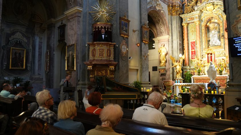 Zdjęcie przedstawia wnętrze kościoła podczas uroczystości. W centrum kadru, na bogato zdobionej ambonie, przemawia mężczyzna w czarnym garniturze z muszką. Po lewej stronie mężczyzna w szarym garniturze gra na flecie, stojąc przy mikrofonie, a przed nim siedzi kobieta grająca na klawesynie. W tle widoczny jest ołtarz z bogatymi złotymi dekoracjami, figurami aniołów i obrazami religijnymi. W ławkach siedzą wierni, uważnie obserwujący przebieg ceremonii, a przed ołtarzem widać zapalone świece, które dodają atmosferze ciepła i uroczystego charakteru.