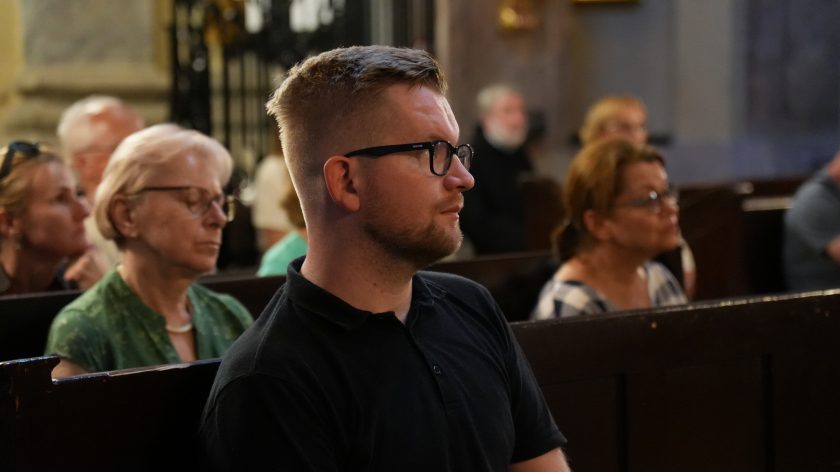 Zbliżenie na mężczyznę siedzącego w ławce kościelnej, który w skupieniu uczestniczy w uroczystości. Mężczyzna ma krótkie blond włosy, nosi okulary i jest ubrany w ciemną koszulkę polo. W tle widoczni są inni wierni, także siedzący w ławkach, część z nich ma zamknięte oczy, co sugeruje, że mogą być pogrążeni w modlitwie lub refleksji.