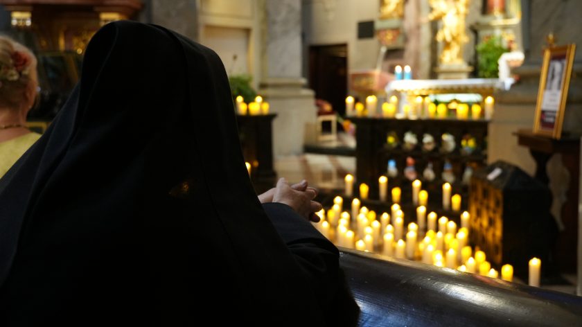 Zdjęcie przedstawia osobę w habicie zakonnym, siedzącą w ławce kościelnej i zwróconą w stronę ołtarza. Osoba ma splecione dłonie i jest pogrążona w modlitwie lub refleksji. Przed nią, na podłodze i ołtarzu, widoczne są liczne zapalone świece, które tworzą ciepłe, złote światło, dodając atmosferze uroczystości nastrojowości i spokoju. W tle widać fragmenty ozdobnego ołtarza oraz inne elementy kościelnego wnętrza.