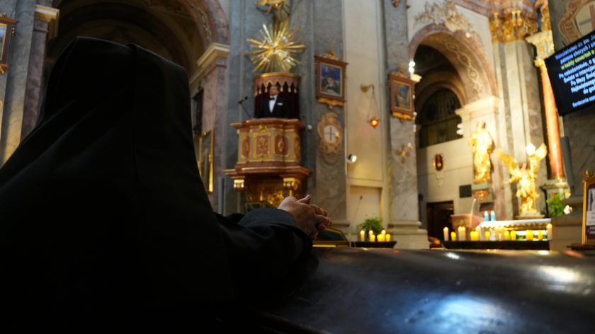 Zbliżenie na splecione dłonie osoby modlącej się w kościele. W tle widoczne są rozmyte, złote światła pochodzące od licznych zapalonych świec, które tworzą ciepłą i nastrojową atmosferę. Zdjęcie oddaje spokój i skupienie osoby pogrążonej w modlitwie.