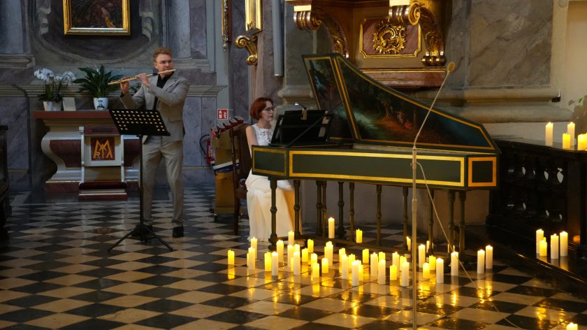 Zdjęcie przedstawia duet muzyczny wykonujący utwór w kościele. Po lewej stronie mężczyzna w szarym garniturze gra na flecie, stojąc przy mikrofonie. Obok niego, po prawej, siedzi kobieta ubrana w białą suknię, grająca na klawesynie ozdobionym malowanym pejzażem. Na podłodze, przed muzykami, ustawione są liczne zapalone świece, które tworzą ciepłą i nastrojową atmosferę. Posadzka jest wyłożona płytkami ułożonymi w wzór szachownicy, a w tle widać fragmenty kościelnego wnętrza z obrazami i elementami dekoracyjnymi.