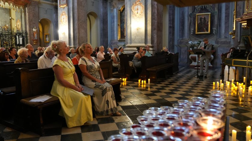 Zdjęcie przedstawia wnętrze kościoła podczas uroczystości. Na pierwszym planie po lewej stronie siedzą dwie kobiety w eleganckich sukniach – jedna w żółtej, druga w białej z czarnym wzorem – zwrócone w stronę ołtarza, skupione na ceremonii. W tle, po prawej stronie, mężczyzna w szarym garniturze gra na flecie, a obok niego kobieta w białej sukni gra na klawesynie. Posadzka kościoła jest wyłożona płytkami ułożonymi w wzór szachownicy, a na podłodze ustawione są liczne zapalone świece, które dodają atmosferze ciepła i uroczystości. W tle widać także innych uczestników siedzących w ławkach, uważnie obserwujących przebieg ceremonii.